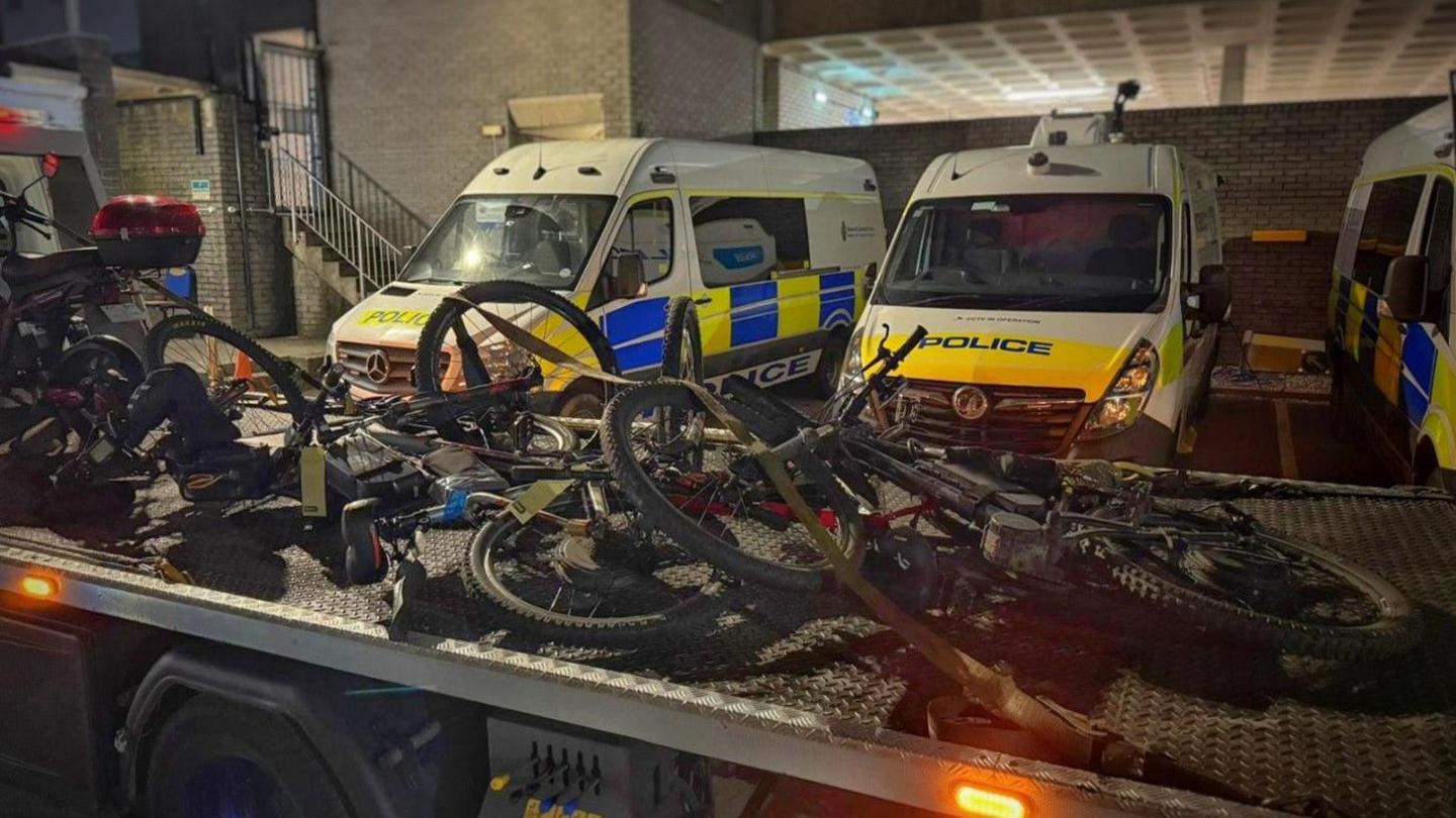 Seized bicycles on the back of a truck with police vehicles in the background. 