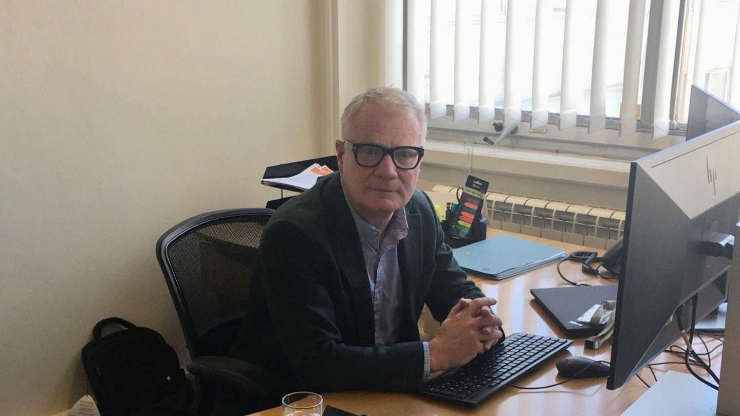 West Midlands mayor Richard Parker with his hands clasped together, wearing glasses facing the camera. He is sitting at a desk with a keyboard and paper and trays are behind him.