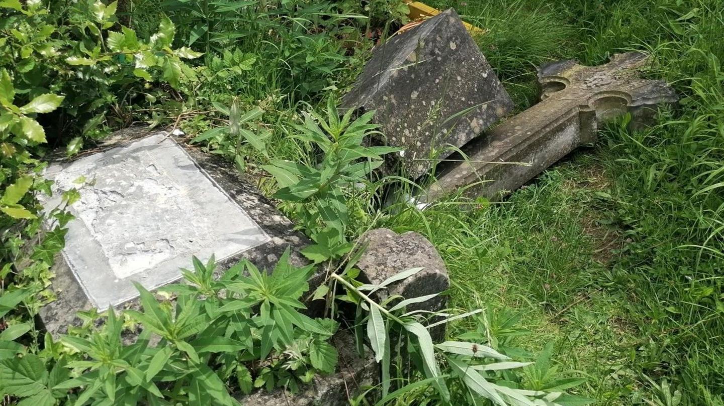 A cross statue which has been knocked off a stone plinth and lays discarded in the overgrown grass beside it.
