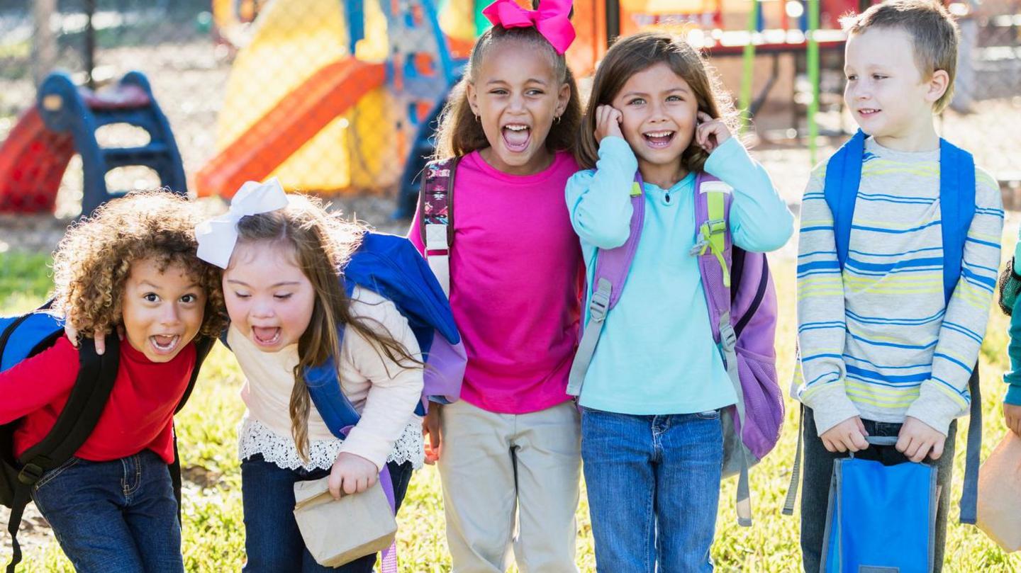 5 children in a row in a sunny park, possibley after school in summer, two are bending down and seem to be shouting in a happy way one has fingers in ears others look on - a happy sunny scene.