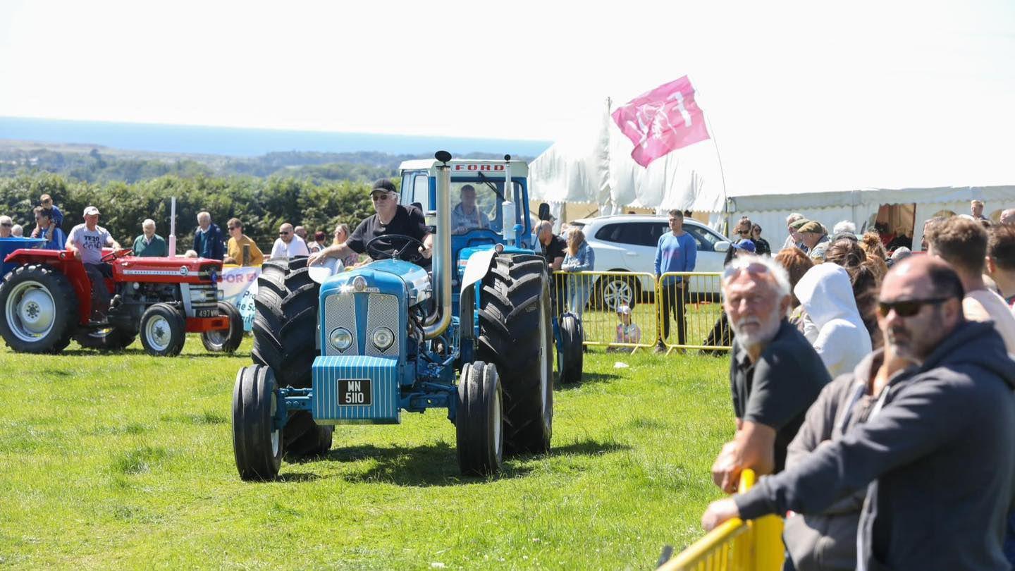 Tractors parading in a field with large crowds watching. The tractor at the front is blue and has an open cab. It is being driven by a man wearing a black T-shirt and baseball cap. 