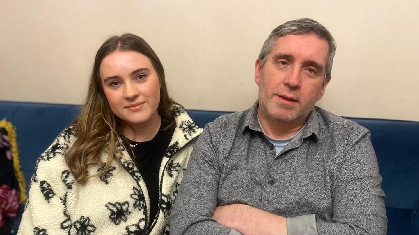 Aimee sitting on a sofa with her dad Damien. Aimee has long brown hair with blonde ends, she's wearing a black top and a cream zip up cardigan with black flowers. Damien has light grey short hair and is wearing a grey shirt with a light blue top underneath.