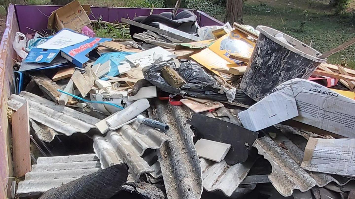 A large amount of waste, including a bucket, roof tiles, piping, cardboard, a car seat, and a plastic bag in a large purple skip in a cemetery