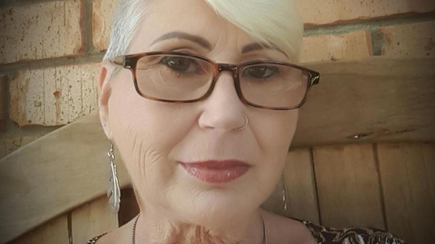 Head shot photograph of a woman with white short hair who is smiling at the camera, She is wearing tortoiseshell reading glasses, has a nose ring and long silver leaf shaped earrings. In the background can be seen the back of a wooden bench and a brick wall.