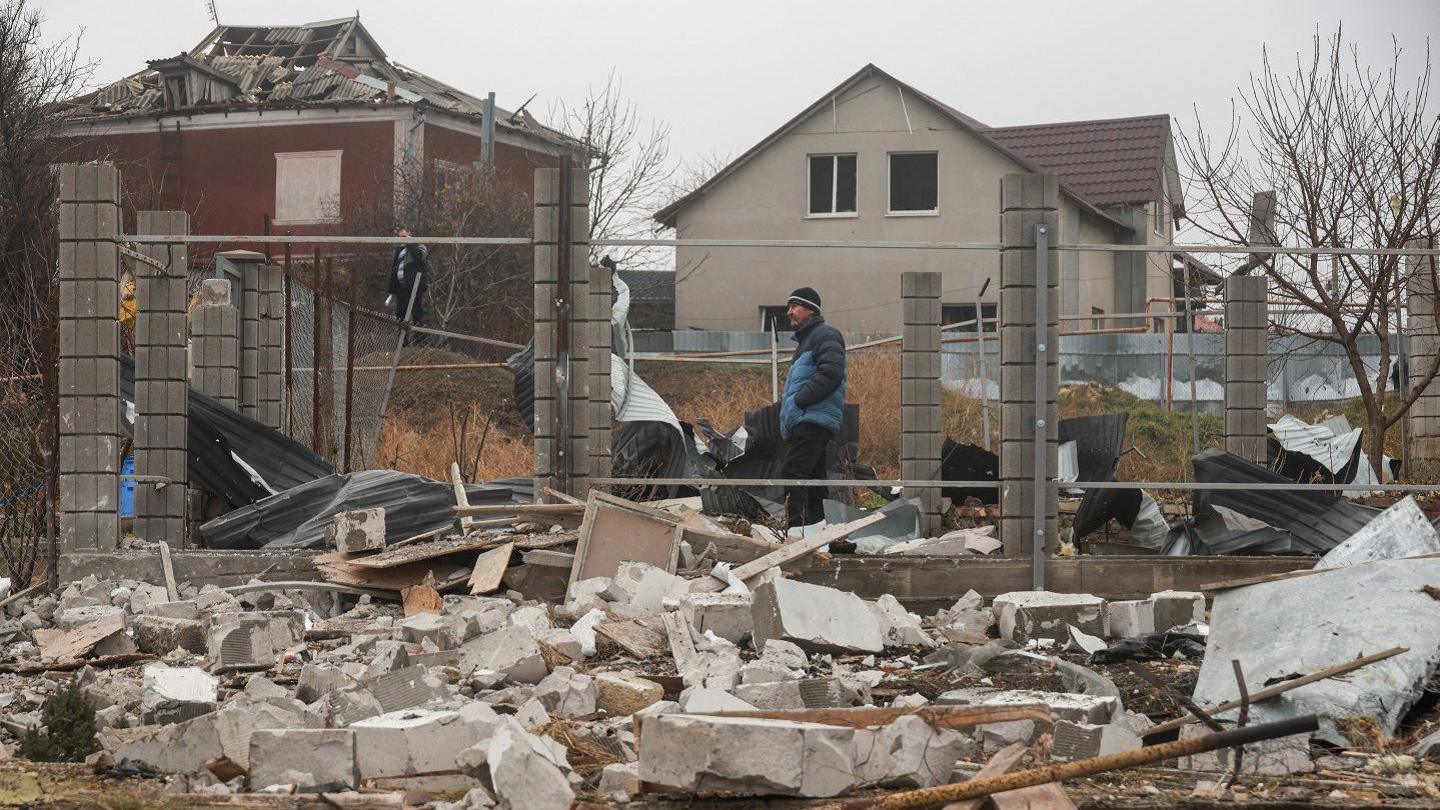 Man standing next to a damaged home. There is rubble in front of him and a what looks to be an unscathed building behind him