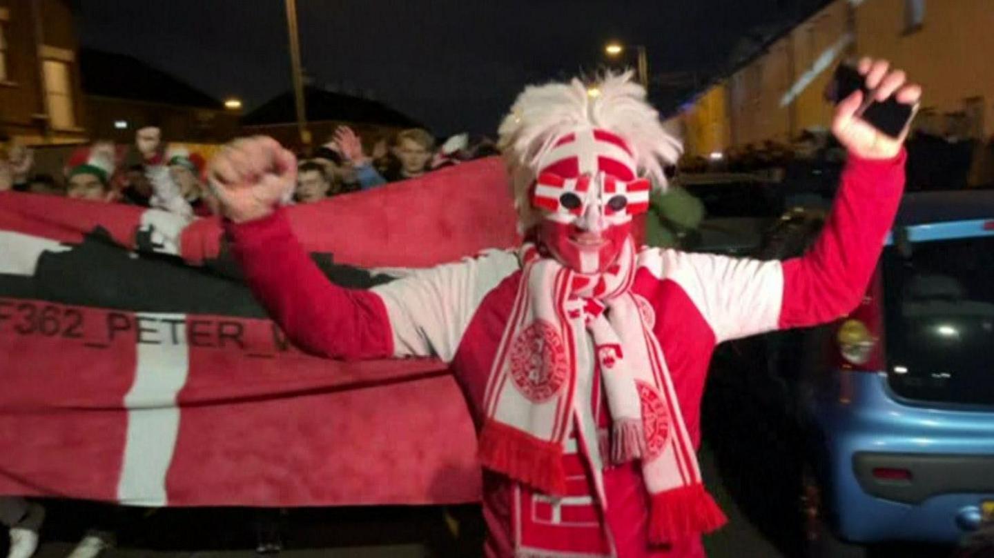 Danish football fans taking part in a parade through Belfast city centre to Windsor Park stadium in November 2023.  One man in the foreground of the photo is holding his arms aloft in a cheering gesture.  He is dressed in his team's colours and his face is covered with red and white mask. 