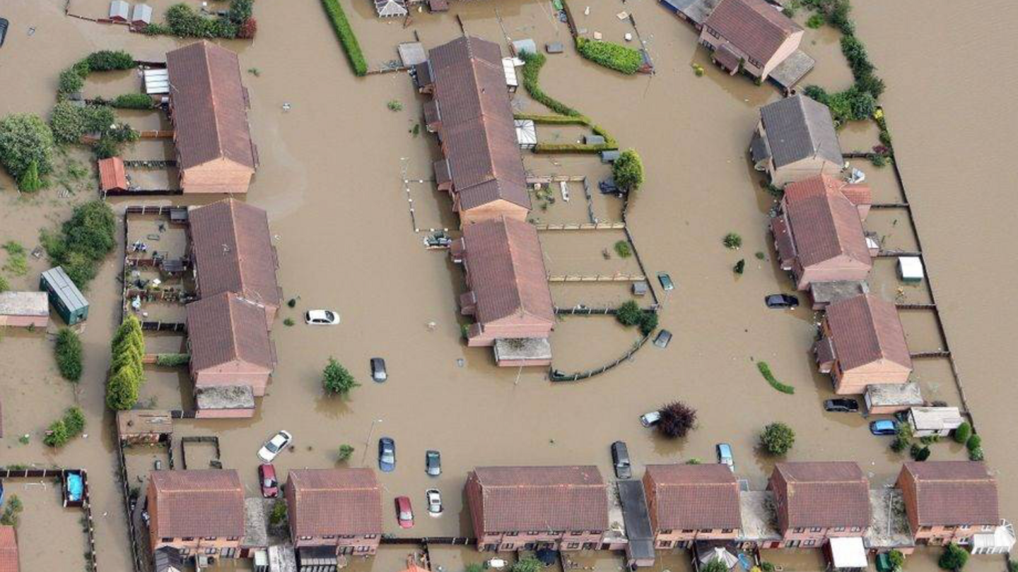 An aerial view of a housing estate whose roads and gardens are submerged under brown flood water. Several parked cars are visible in the flooded streets.