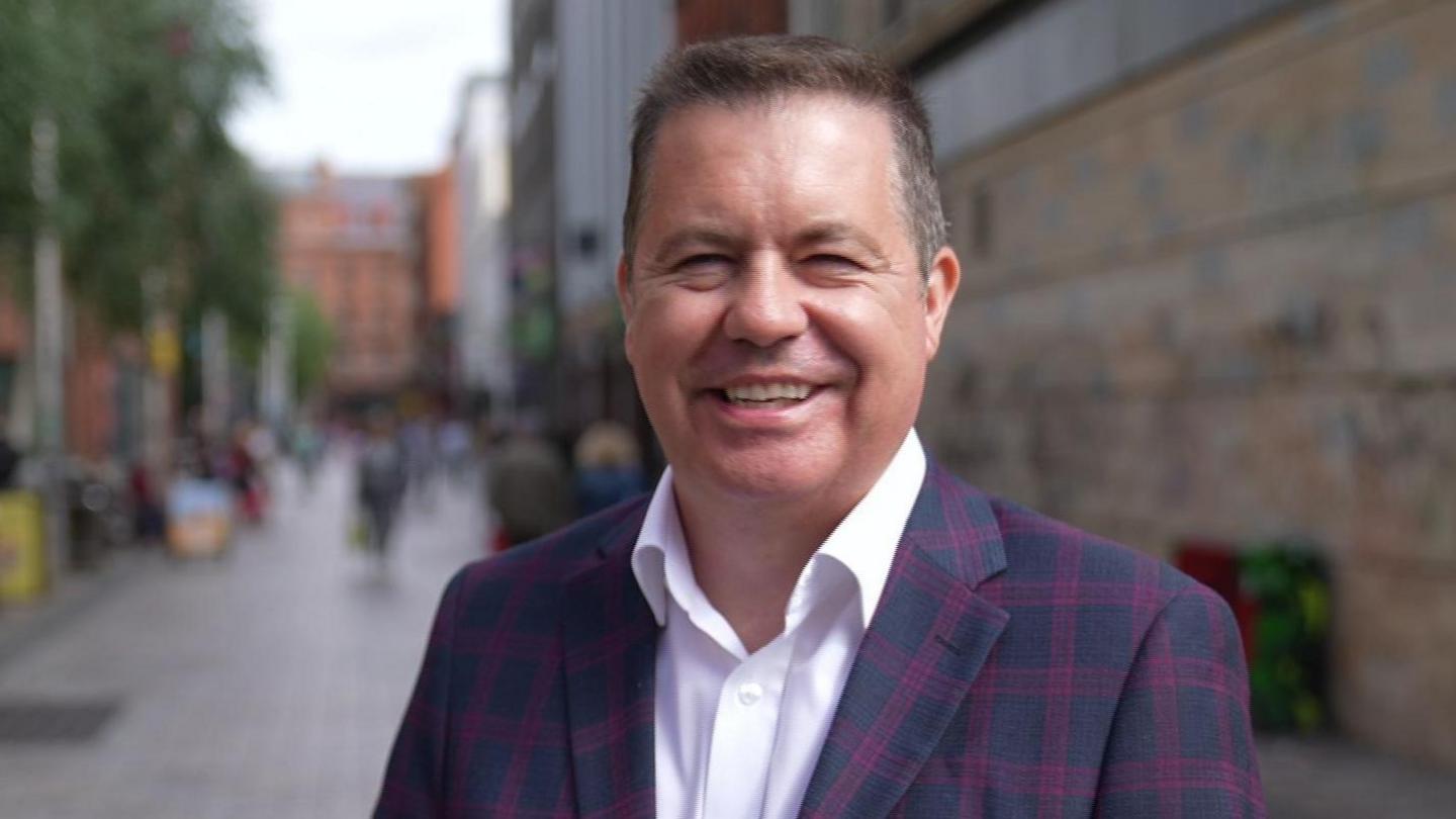 Glyn Roberts, chief executive of the Retail NI, smiling wearing a white shirt and checked navy and purple blazer.