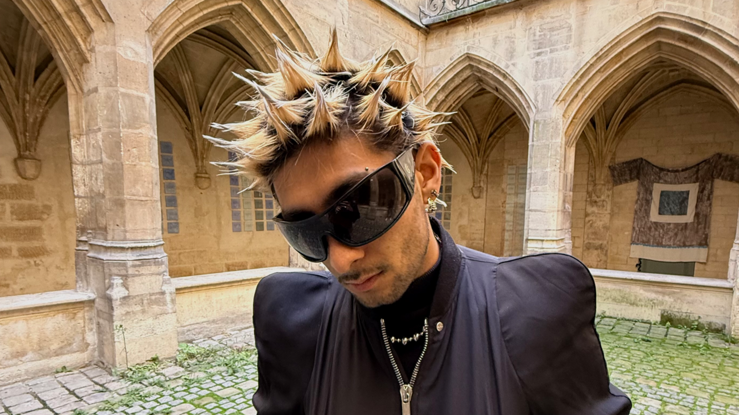 Aly Meghani is looking down, wearing huge black sunglasses and a black jacket. His hair is in blonde spikes.