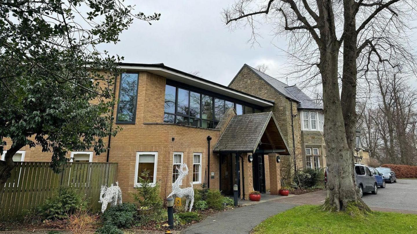 St Cuthbert's Hospice is a brick building with an archway above the entrance. The windows on the ground floor have white frames, while those above have black frames and tinted glass. A large tree is growing at the front of the property close to where three cars are parked.
