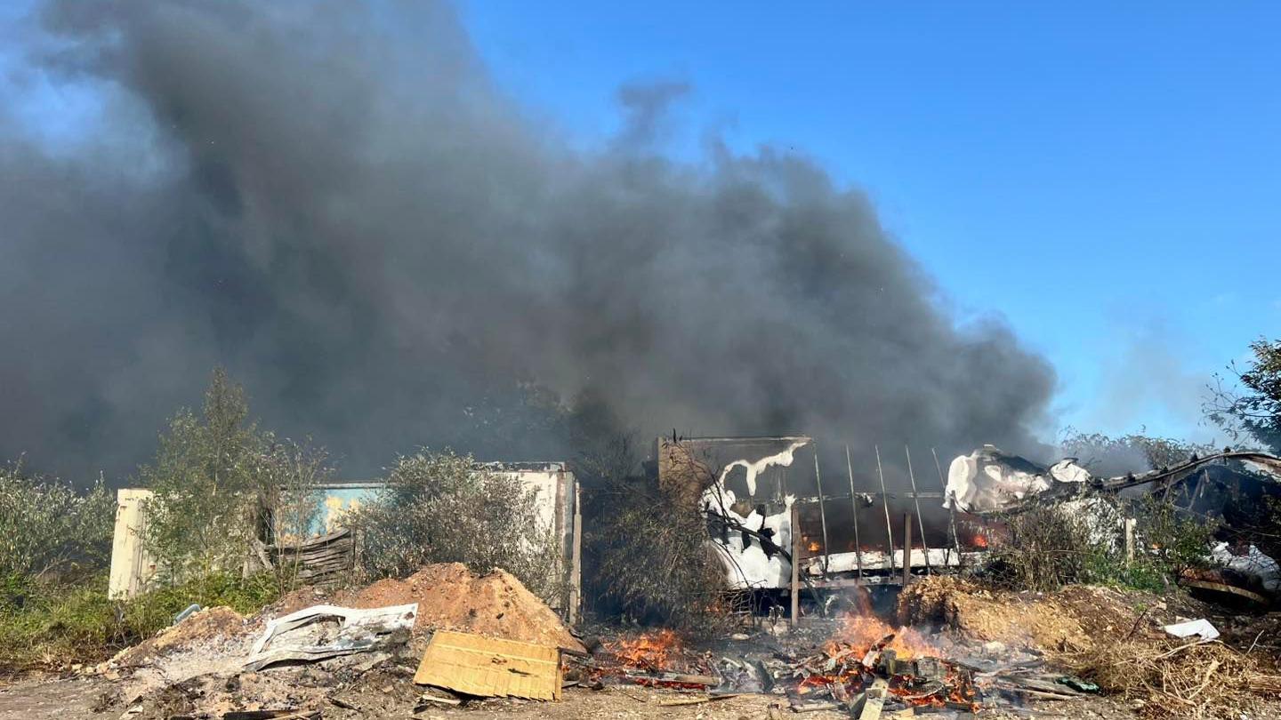 A pile of burning rubbish and sheds surrounded by thick black smoke which is filling the sky
