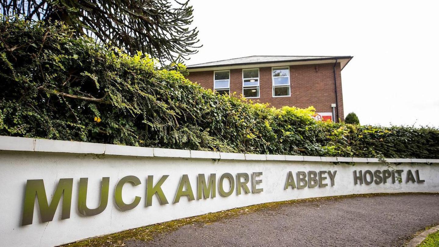 A sign says Muckamore Abbey Hospital on a low white wall in front of bushes and a red-brick building