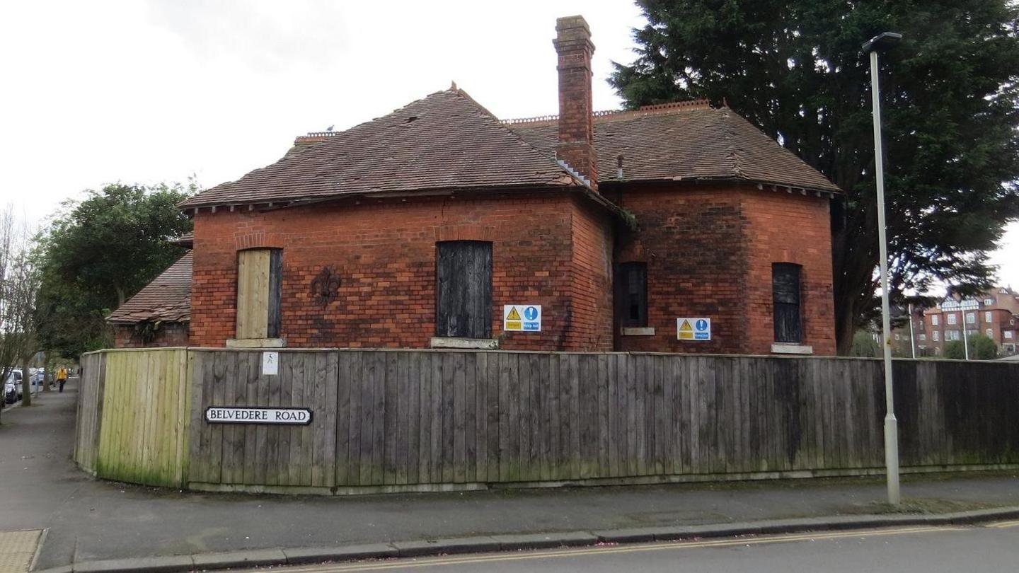 The back of a dilapidated Tudor-style wood and brick building. It has blue and yellow signs on it with safety warnings. It is viewed from the road. A street sign says Belvedere Road.