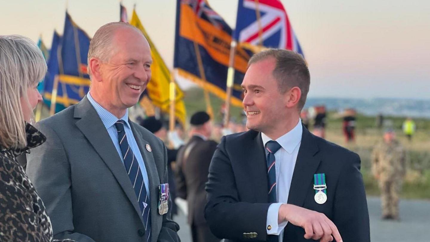 Steven Cartwright in a dark blue suit speaking to the Lieutenant Governor Jerry Kyd who is wearing a grey suit. Flags and people in military uniforms are in the background. 