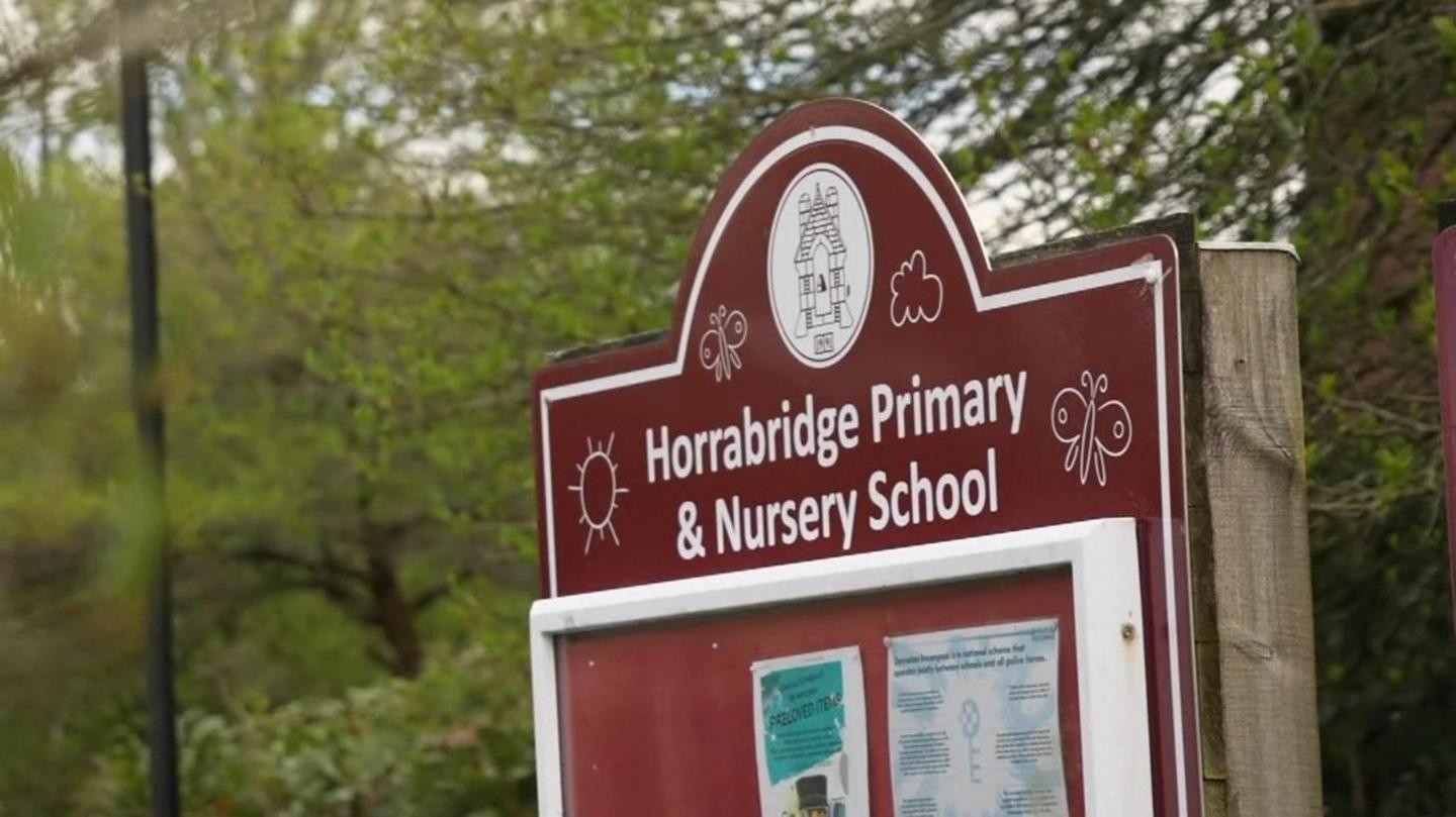 A burgundy sign which has the words 'Horrabridge Primary and Nursery School'. There is posters below it, and trees in the background.