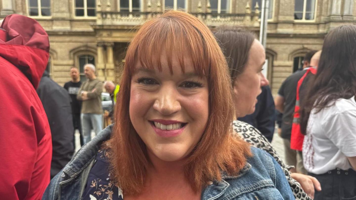 Woman with shoulder length red hair, patterned top and denim jacket. She is stood in front of a crowd of people.