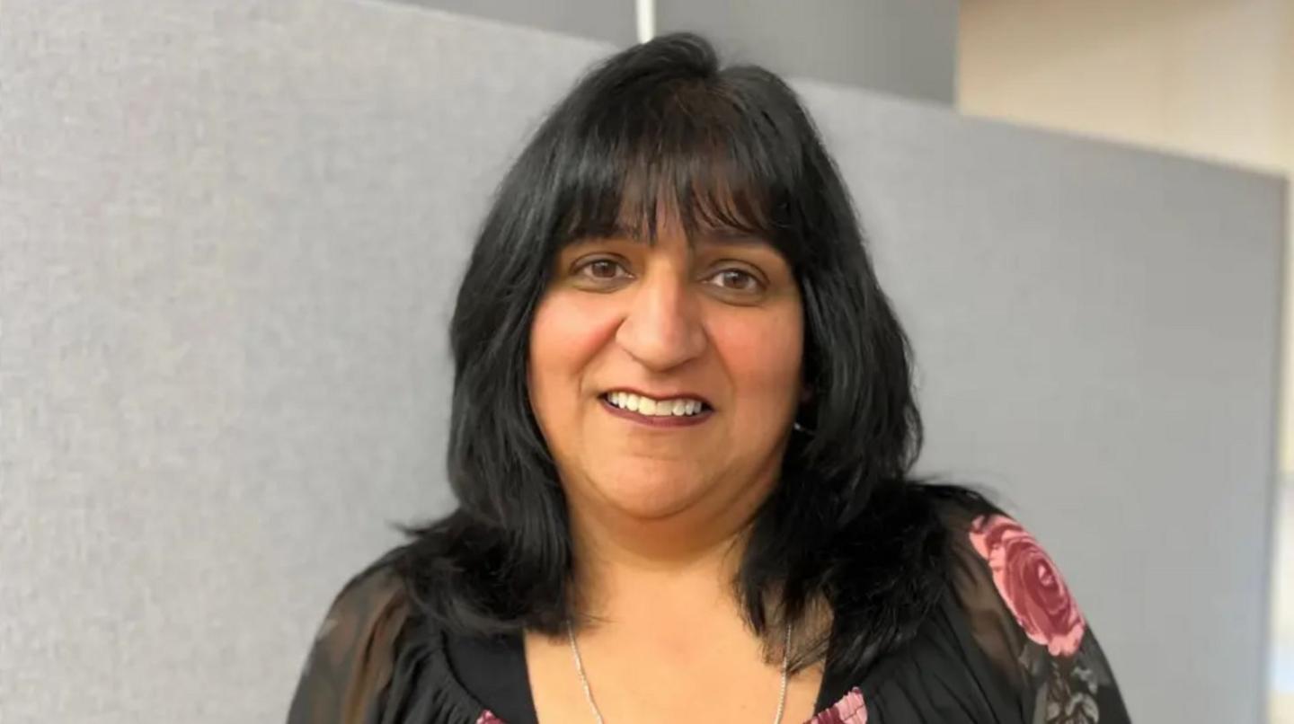A woman with long black hair and a black top with a pink floral pattern on it in a room with a grey wall