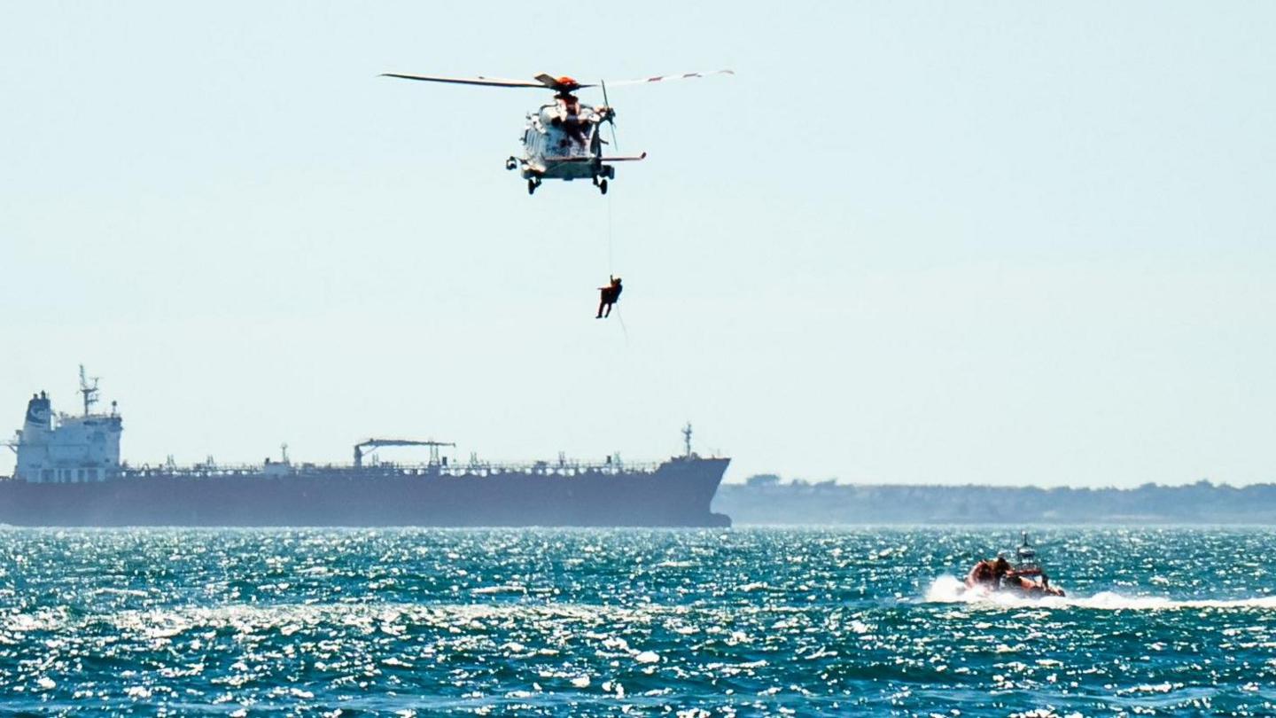 A person hanging below a helicopter hovering over the sea with a lifeboat below. In the background is a large bulk carrier ship