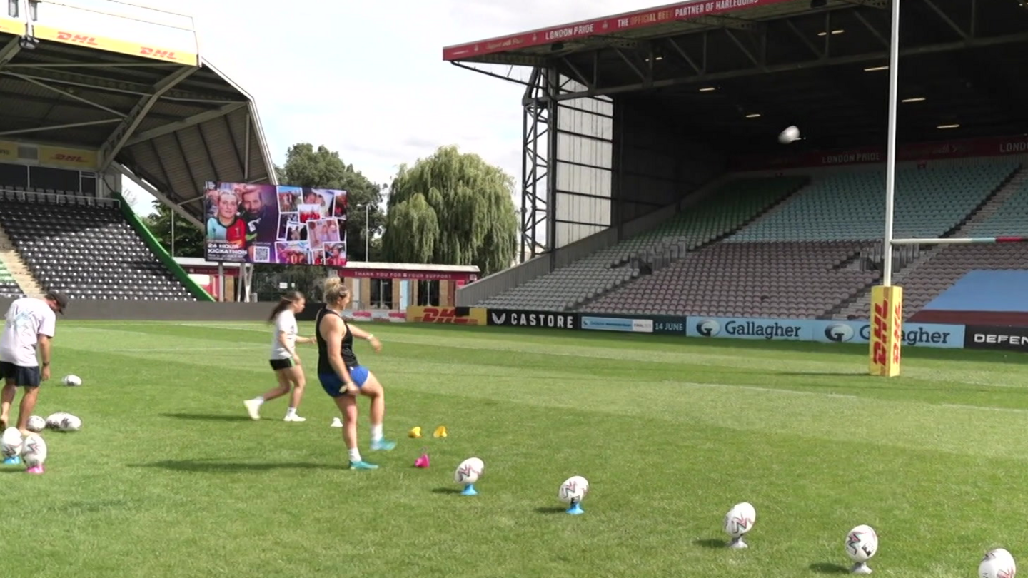 Ella Cromack, wearing a dark T-shirt and blue shorts and light blue boots, kicking a rugby ball. Other rugby balls are lined up ready for her to kick.