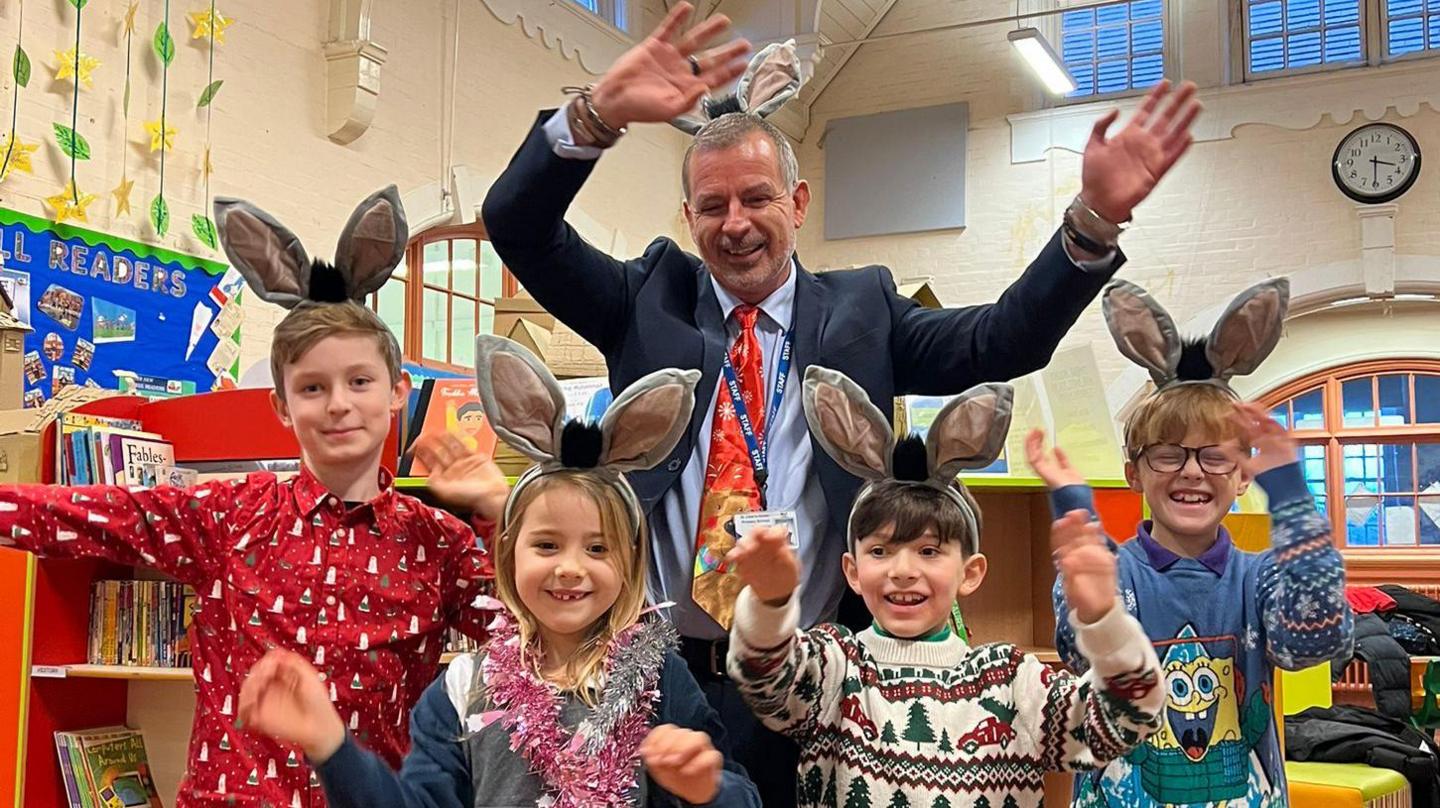 Simon Billings waving his hands in the air while wearing a navy suit jacket, blue shirt and red tie and smiling. In front of him are three boys and a girl, all who are wearing Christmas clothing and smiling. All pictured have donkey ears on their head and are standing in what looks like the school library.