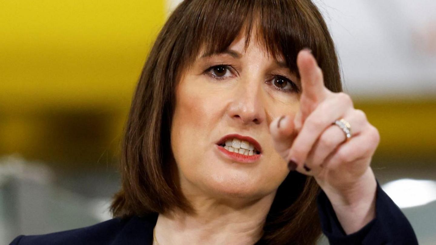 Rachel Reeves has dark brown hair cut into a bob with a fringe. She is wearing red lipstick, a black jacket and is pointing at the camera. She is wearing dark nail varnish and has two rings on her ring finger.