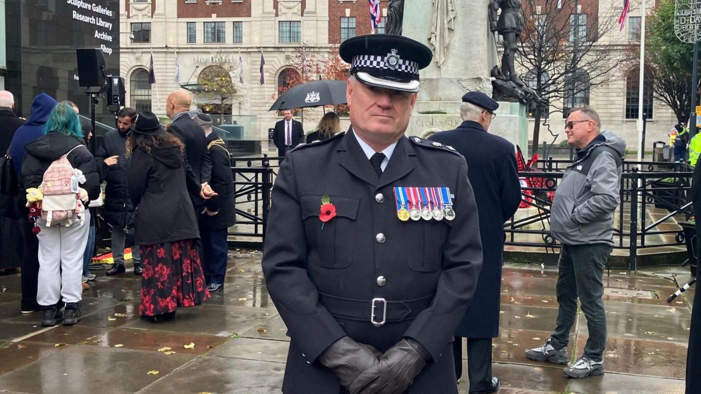 A uniformed police officer stands facing the camera. He is wearing a cap, dark gloves and a dark uniform with several medals on the chest and a red poppy pin. Behind him are a group of people gathered near a tall stone war memorial. The memorial features a bronze statue of a soldier with several Union Jack flags visible.