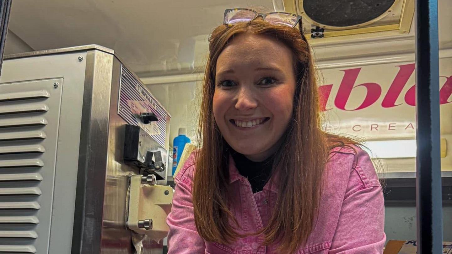 Lauren Evans leaning out of ice cream shop window wearing a pink shirt. She has red long hair and glasses pushed up into her hair