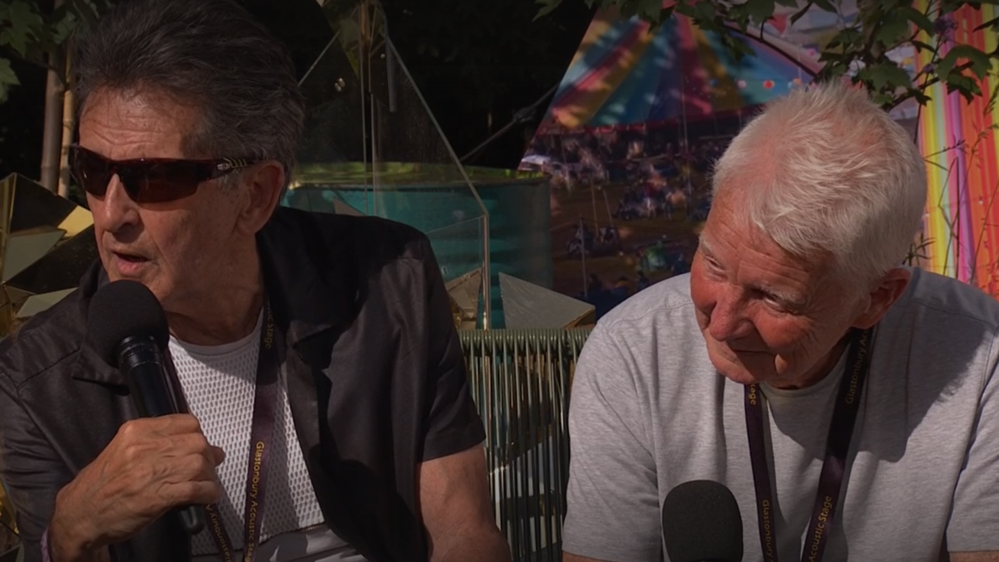 Frank Allen and John McNally sit in a garden at Glastonbury holding microphones.