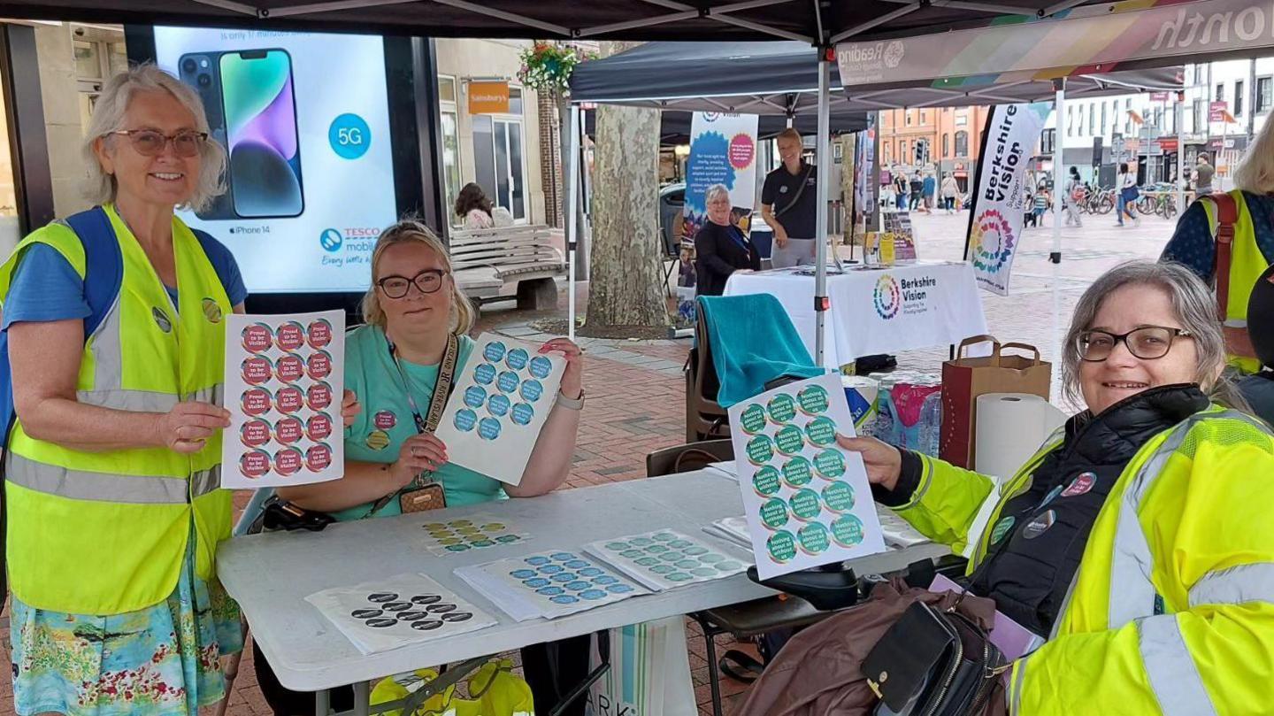 Three women wearing high vis jackets are crowded around a white table. Two are sitting and one is standing. They are holding up pages of red, blue and green stickers. There are more stickers on the table.