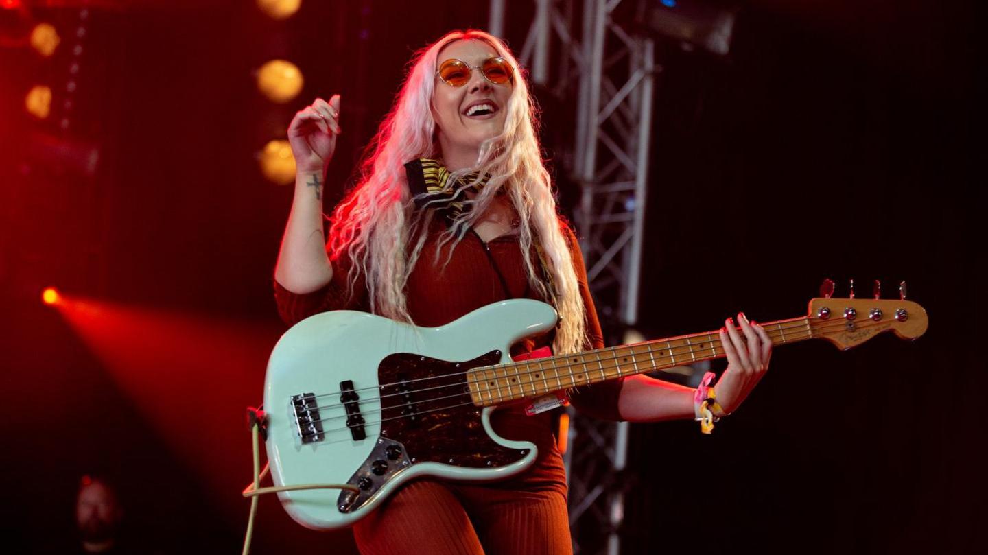 Katie Lloyd of the Lottery Winners wearing a red outfit and playing a white bass guitar