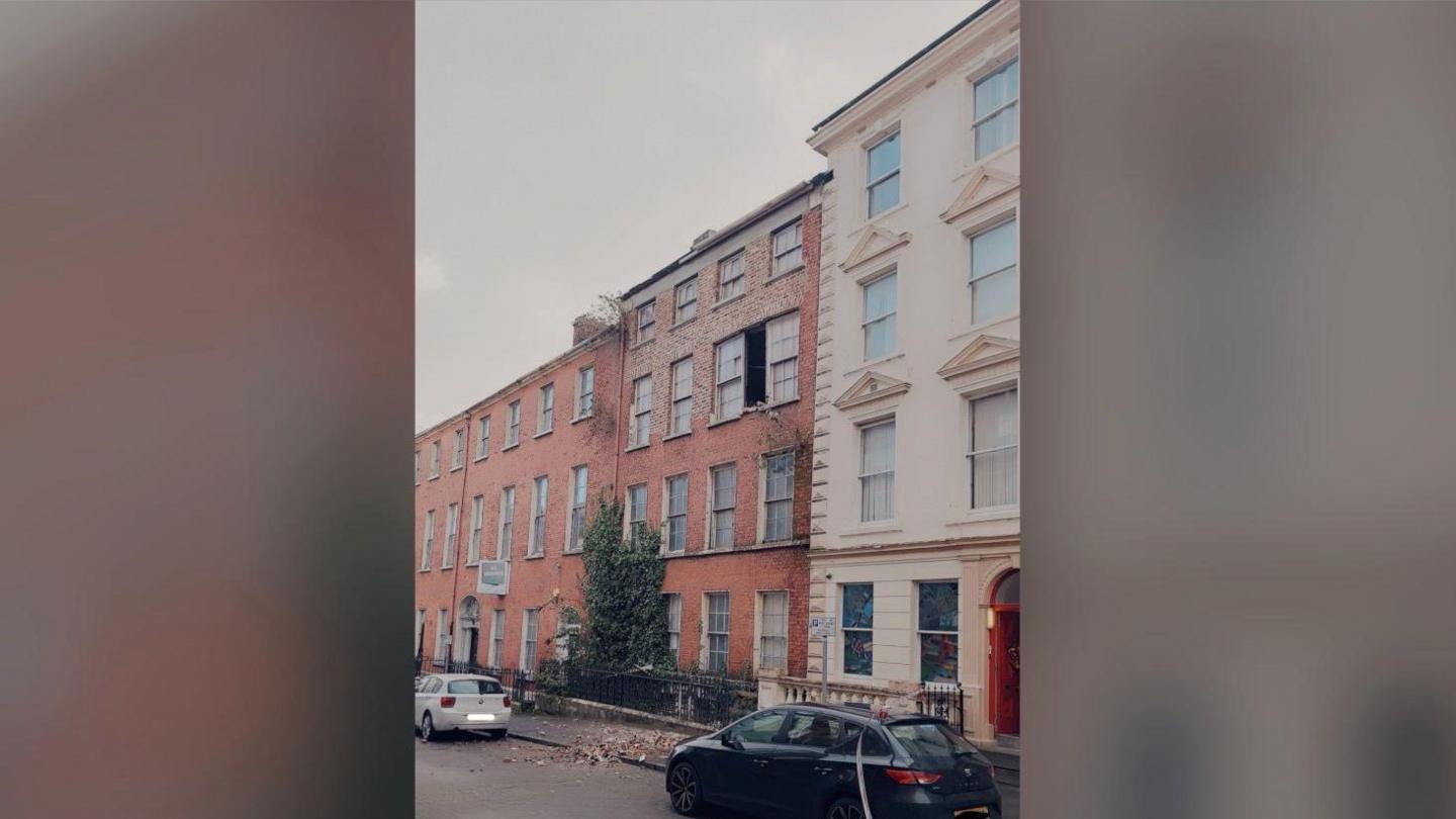 A wider view of terraced buildings on Pump Street in Derry.  There is a gap between two second-storey windows at the front of a red-bricked building in the middle of the photo.  There is a pile of red bricks on the pavement below. 