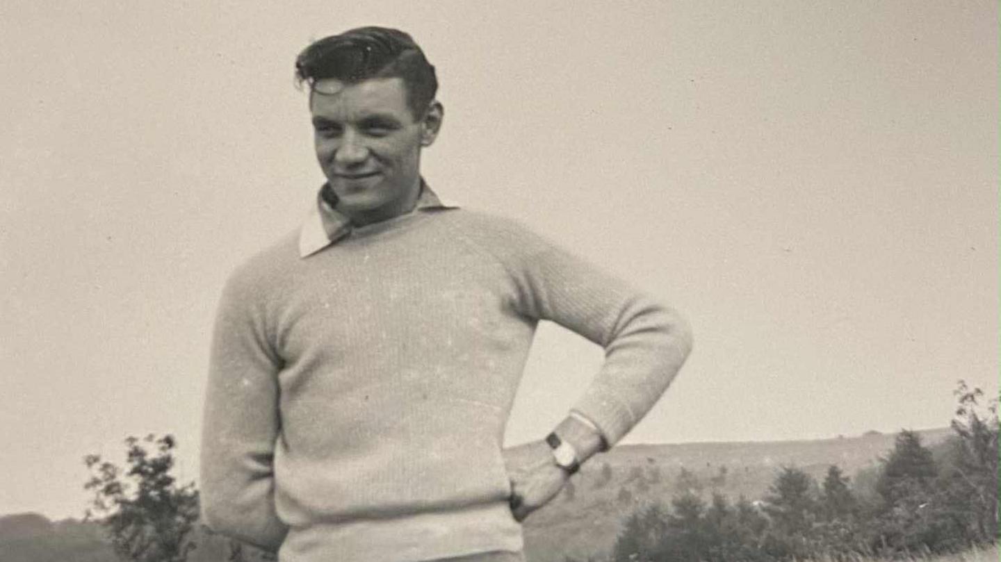 A black and white photo of a man standing in a field with hills and trees in the background. He wears a light jumper over a collared shirt and light trousers, with one hand on his hip. A watch peeks out on his wrist, he has dark hair and looks away from the camera to the left.