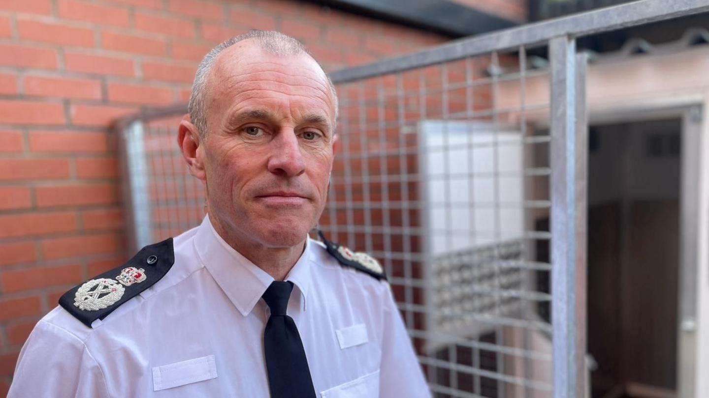 Chief Constable Mark Hobrough, who has closely cropped hair and is wearing a white shirt with epaulettes and black tie, standing in front of a brick wall and what appears to be a security gate, with a small temporary building behind the gate.