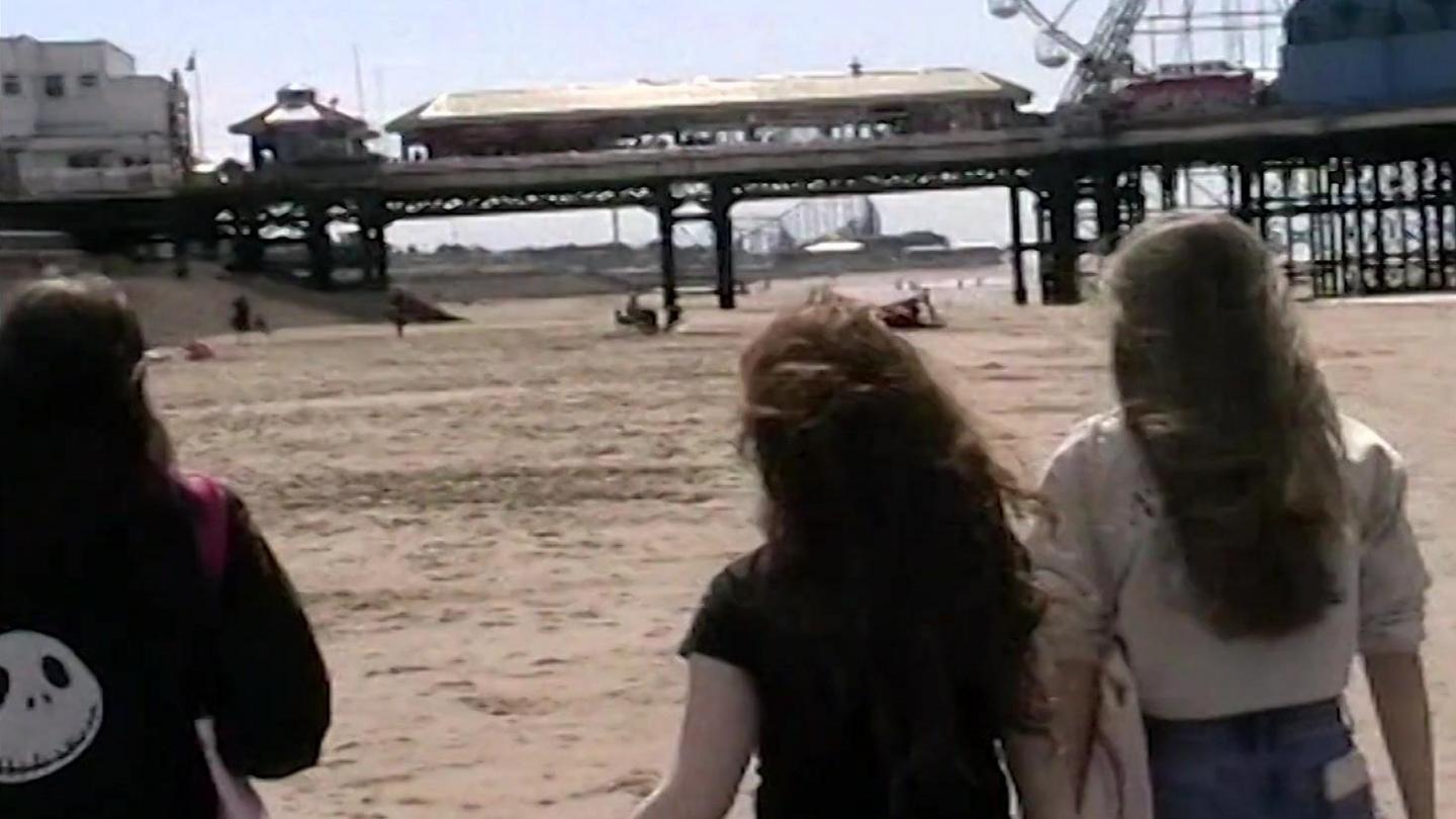 Three teenaged girls, all with long dark hair and with their backs to the camera, walk along the beach towards Blackpool Pier.