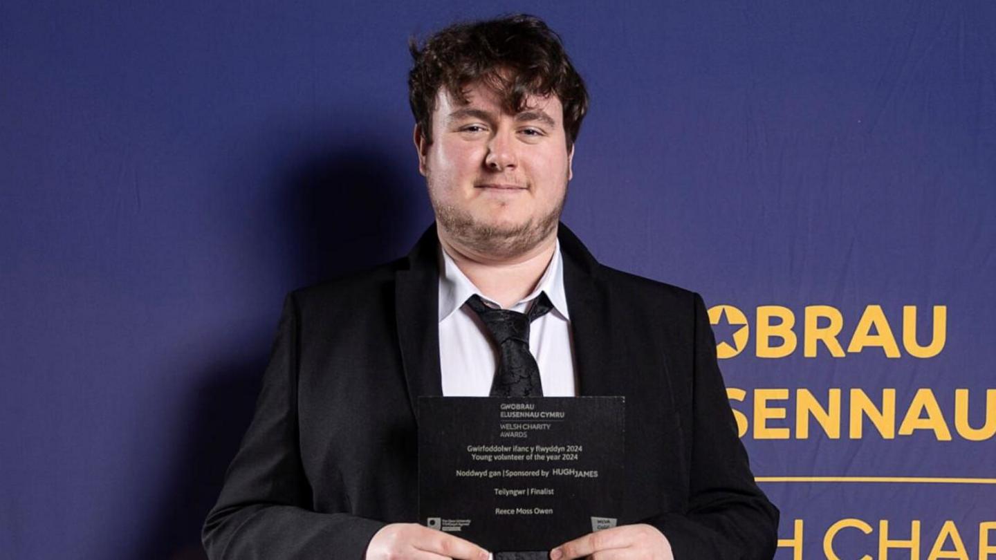 Reece yn gwisgo siaced smart ddu gyda crys gwyn a tei du. Mae'n dal gwobr