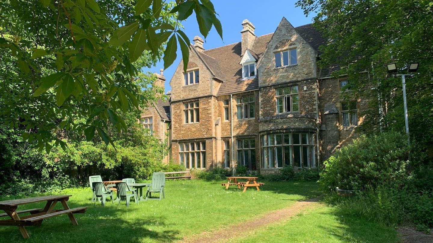 The Garden House - a large old stone building with large windows and a lawned area in front. There are brown wooden benches and green chairs and a table.