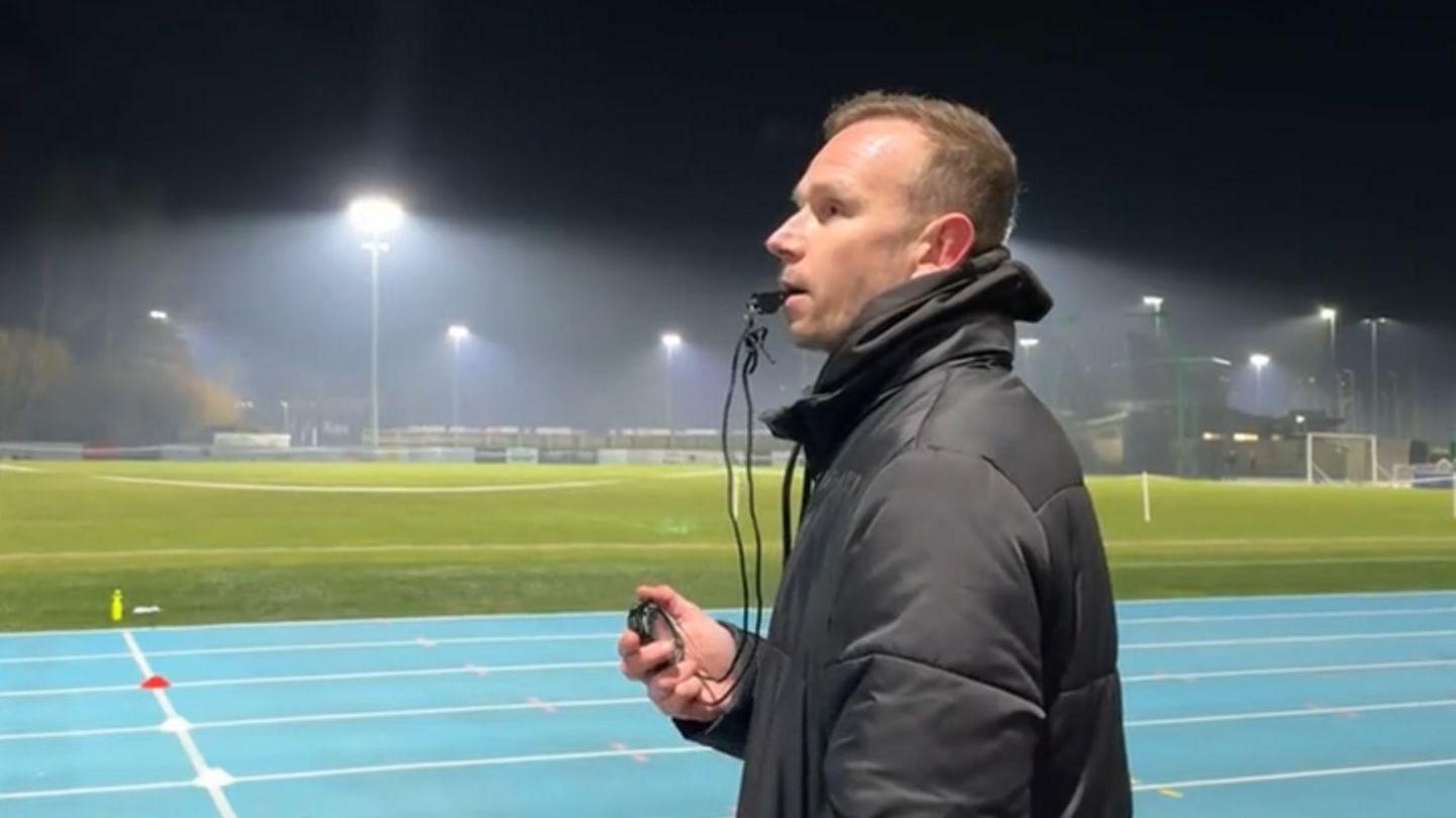 A picture of Tom Druce with a whistle in his mouth and a stopwatch in his hand. He is standing in front of the athletics track.