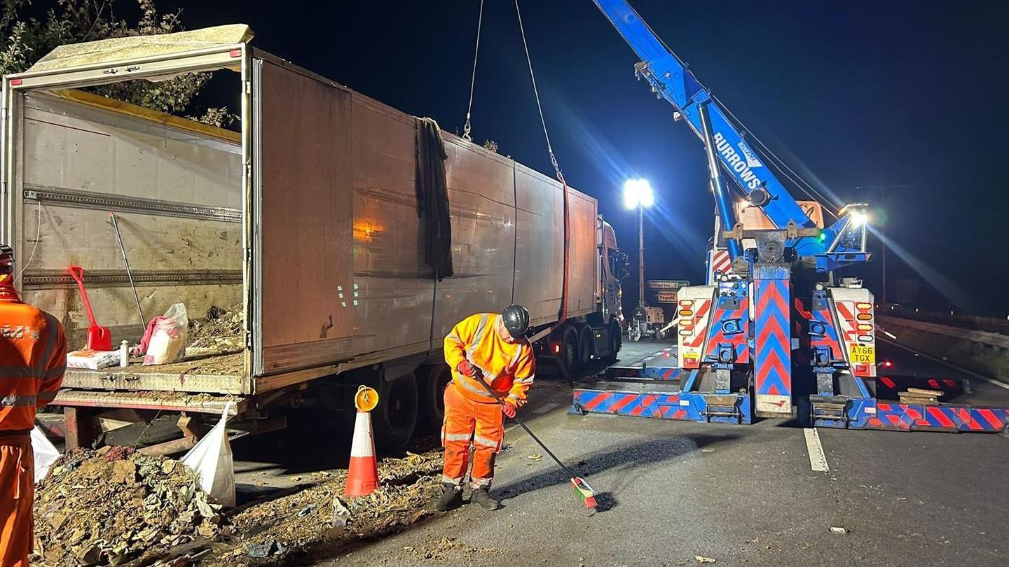 A large white trailer full of waste at the side of a layby has been emptied, with a few bags and a red spade still inside. A blue crane with ropes is attached to each side of the trailer. A man in an orange hi-vis jacket and a black helmet is sweeping up.