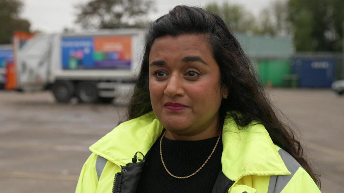 Lynsey Smith, wearing a yellow high-viz coat with black top and a thin gold chain, standing in a council depot with a Newham waste disposal lorry in the background