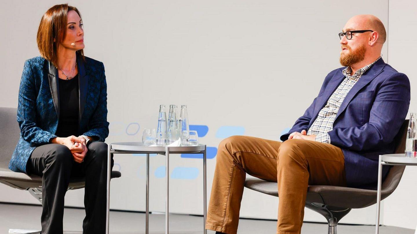 Joel and a woman in conversation on a stage at an event. They both have microphones attached to them and are sat down. 