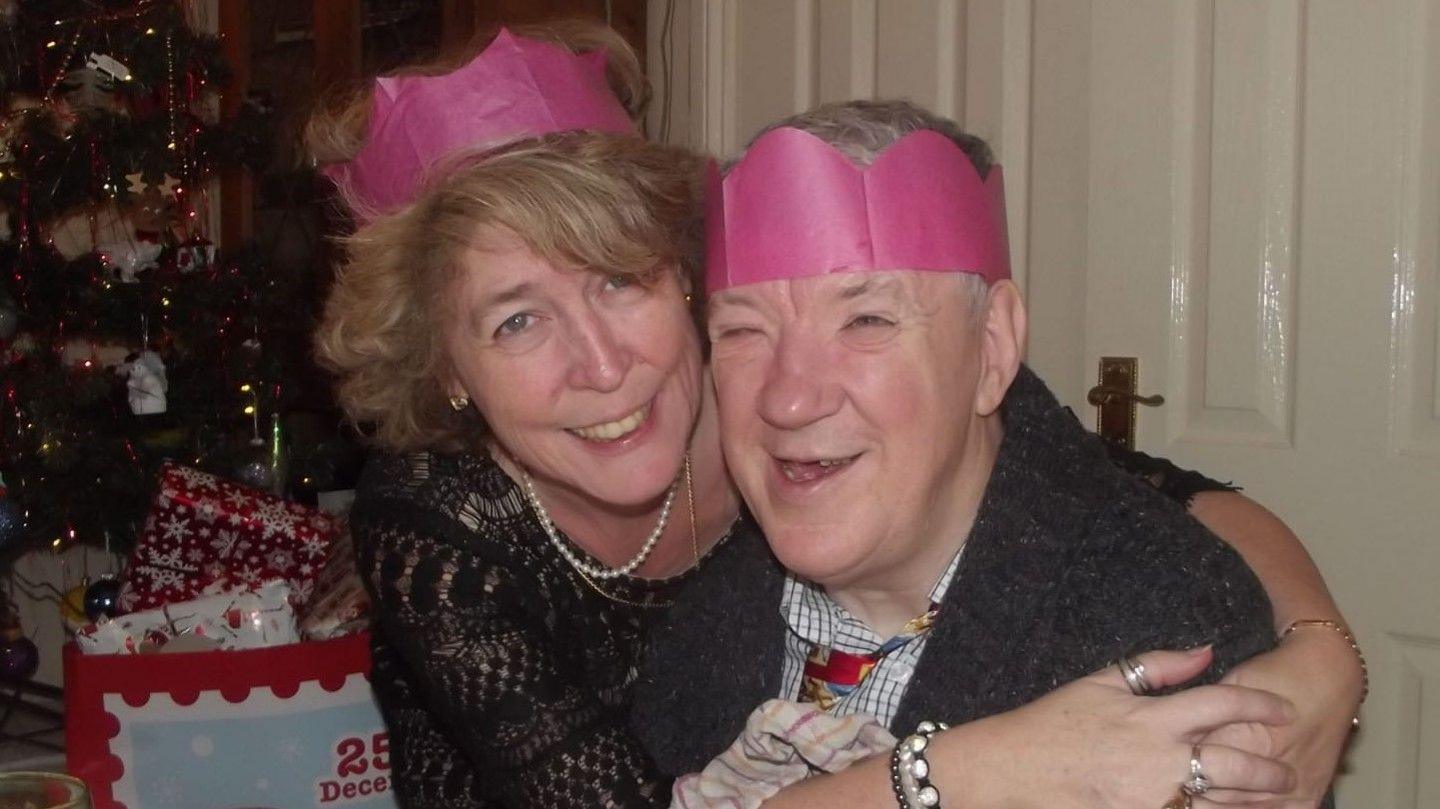 A woman and a man with paper hats on their heads with a Christmas tree in the background. The woman has her arms round the elderly man who is sitting down. They are both smiling. 