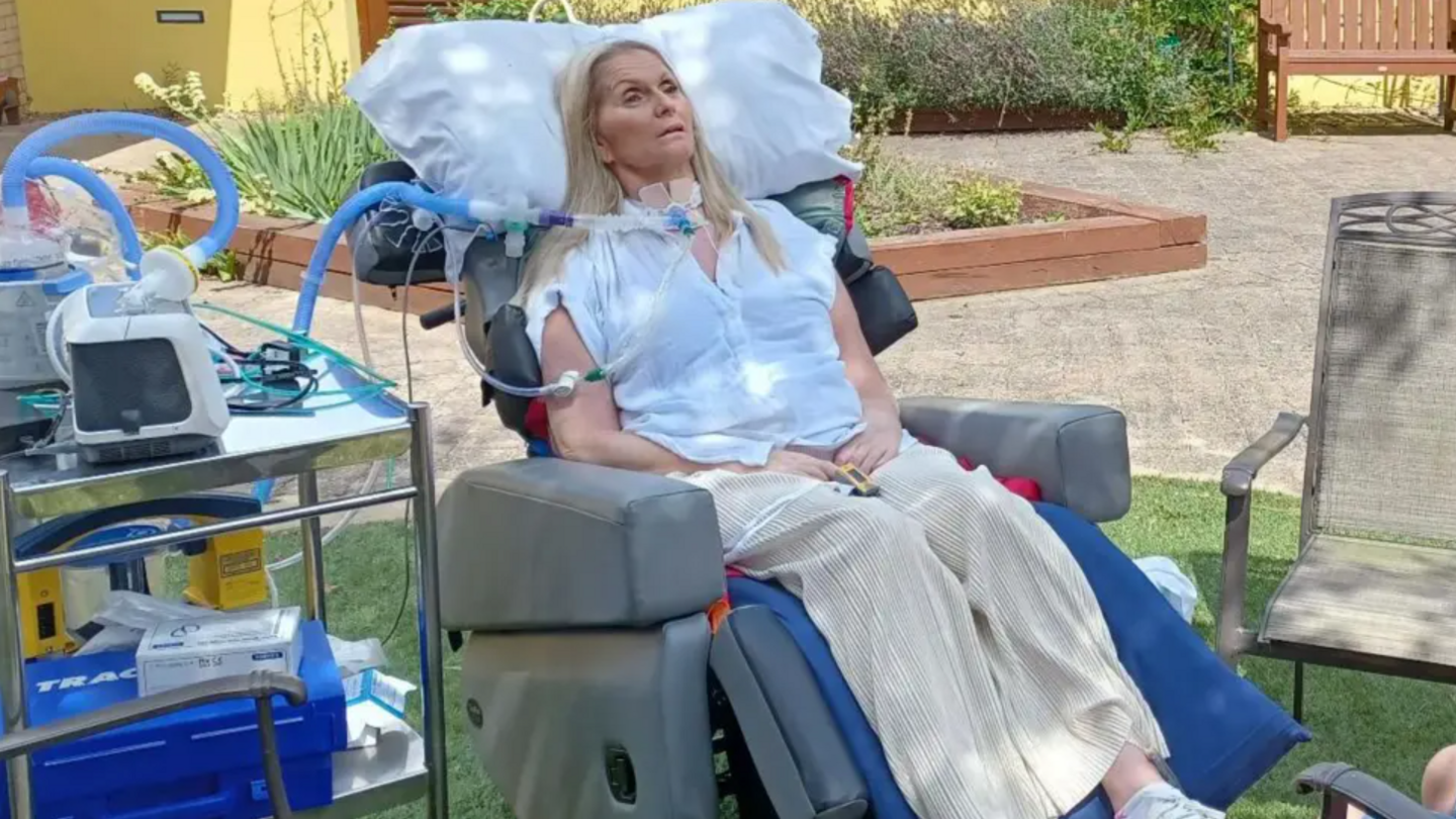 A disabled woman sitting in a specialist chair in a garden. Tubes are connected to her throat. 