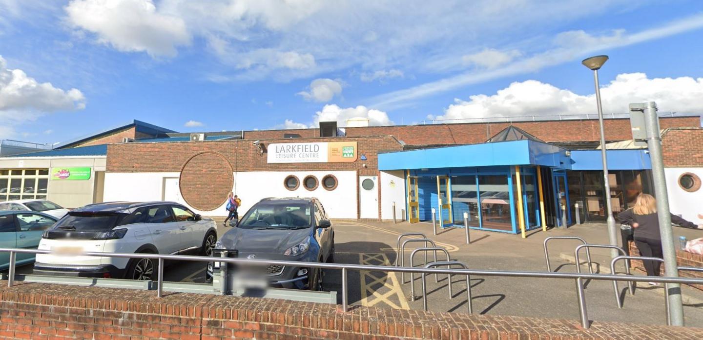 Larkfield Leisure Centre from the outside with a car park in front. The sky is blue with white clouds.