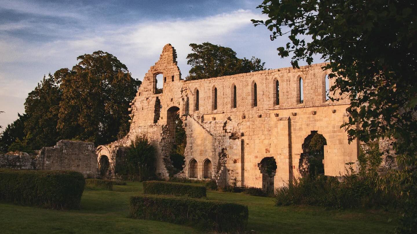 A stone abbey which is in ruins set in gardens with neatly-cut hedges. 
