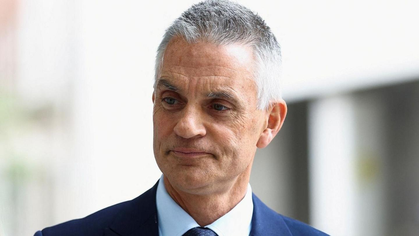 Tim Davie has short grey hair and is wearing a navy suit and light blue shirt