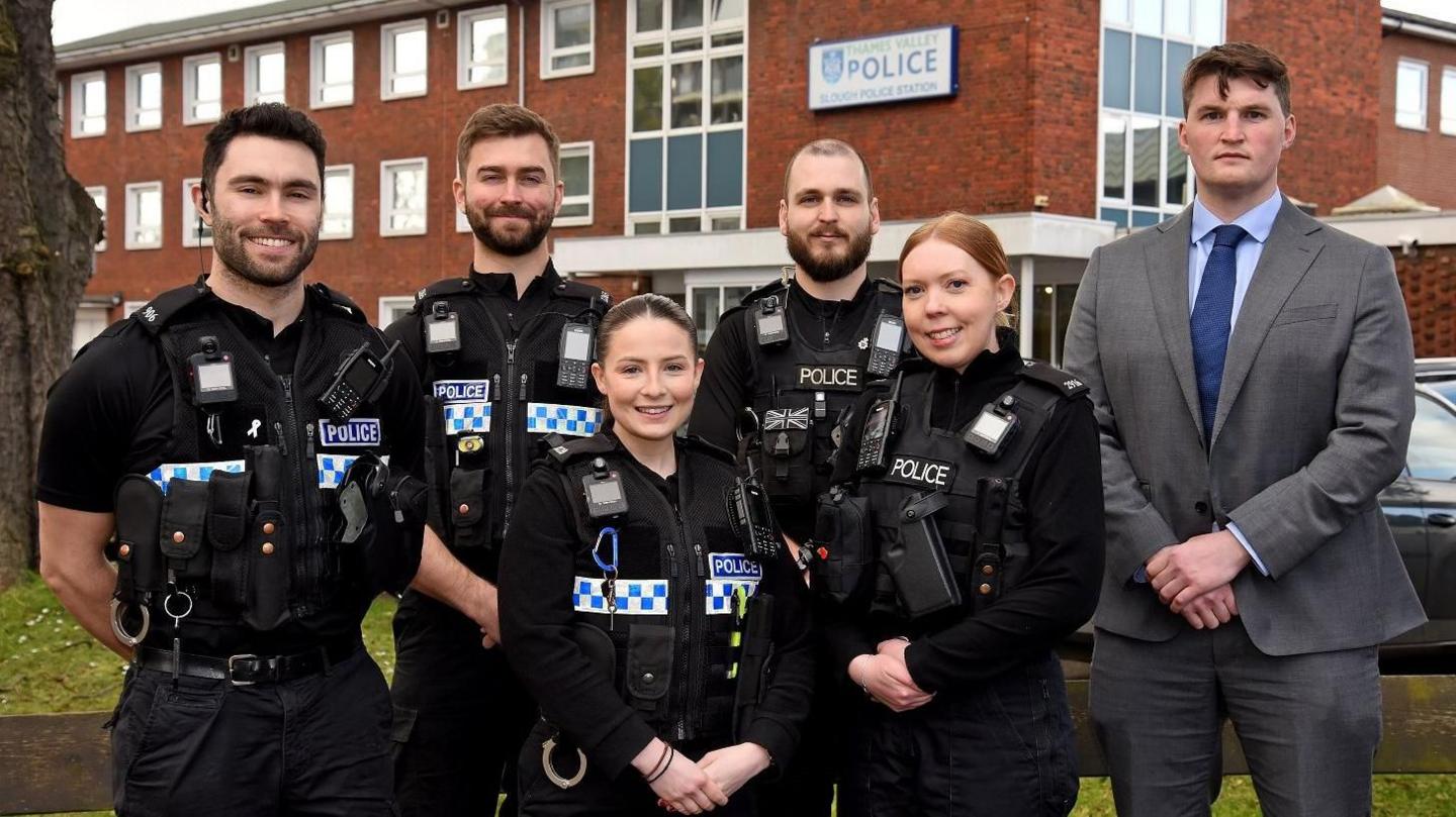 Two women and three men in black police uniform, and a man in a grey suit, standing and smiling in front of a brick building with Thames Valley Police written above the door.