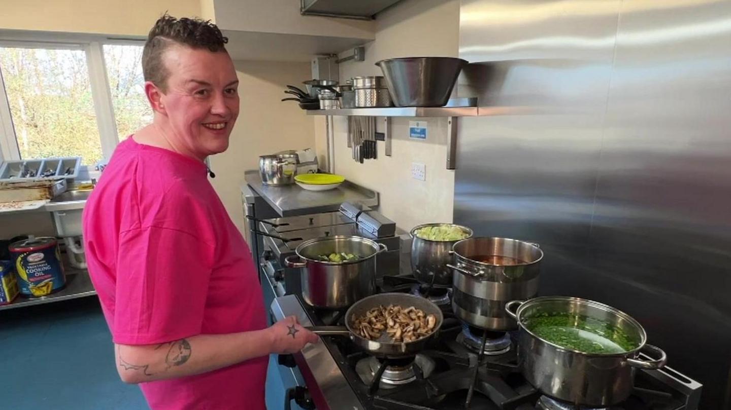 Emma Hall has short dark hair and is wearing a pink T-shirt. She is smiling at the camera as she stands over a hot stove with several pans of food on the hob. She is in a kitchen