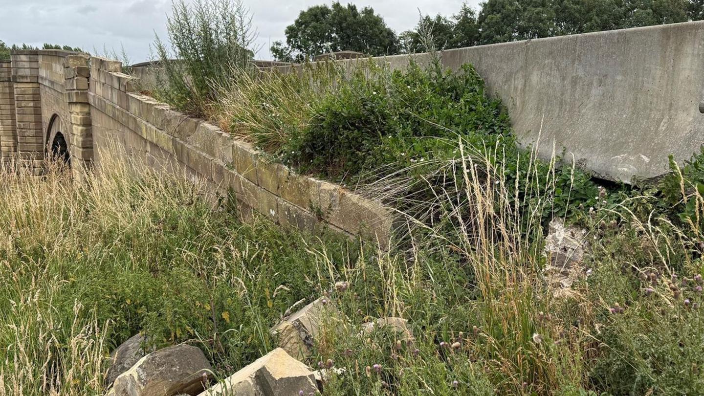Morton-on-Swale bridge is a listed stone bridge over a river in North Yorkshire. The side of the bridge on the approach has temporary concrete barriers due to damage to the older structure.