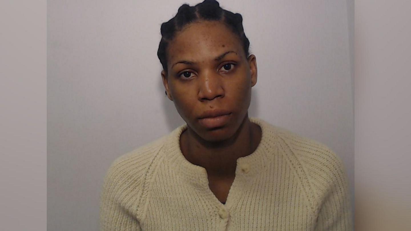 Ruth Auta looks straight at the camera in this police photo. She has braided hair which is tied back and is wearing a cream coloured cardigan. There is a Greater Manchester Police logo in the top left hand corner.