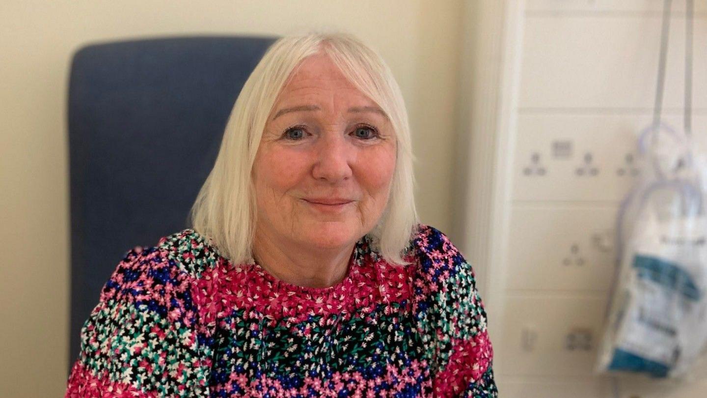 Lesley Harrison is looking into the camera with a small smile. She wears a floral patterned top and is sitting in a hospital chair.